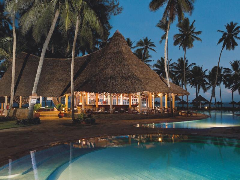 Vista serale di un resort tropicale con piscina illuminata e bar con tetto di paglia circondato da palme.