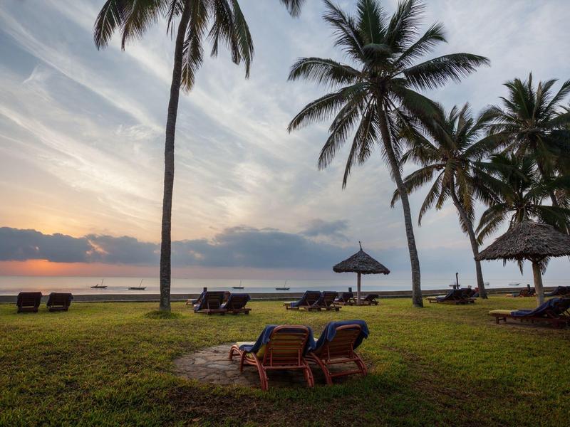 Tramonto sulla spiaggia con palme e sedie a sdraio su un prato verde.