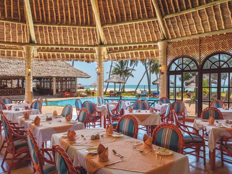 Ristorante con tavoli apparecchiati e vista sulla spiaggia e sul mare sotto un tetto di paglia.