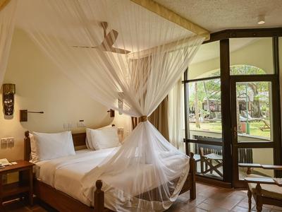 Hotelový pokoj s manželskou postelí, bílou moskytiérou a francouzskými okny s výhledem do zahrady.