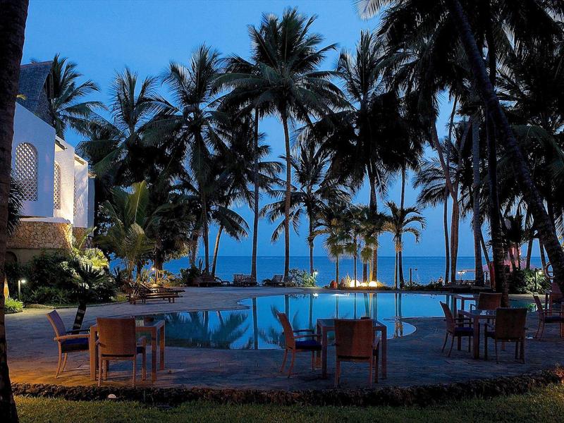 Osvětlený bazén obklopený palmami s výhledem na moře a venkovním nábytkem.