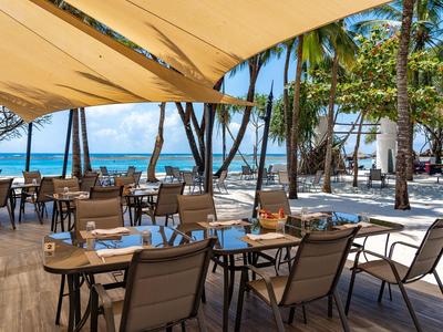 Terasa restaurace na pláži s prostřenými stoly, výhledem na oceán a palmy.