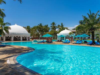 Velký bazén s modrými lehátky, palmami a slunečníky pod jasnou oblohou.