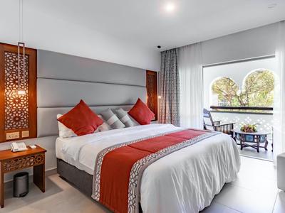 Pohodlný hotelový pokoj s velkou postelí, červenými akcenty a balkonem s výhledem na stromy.