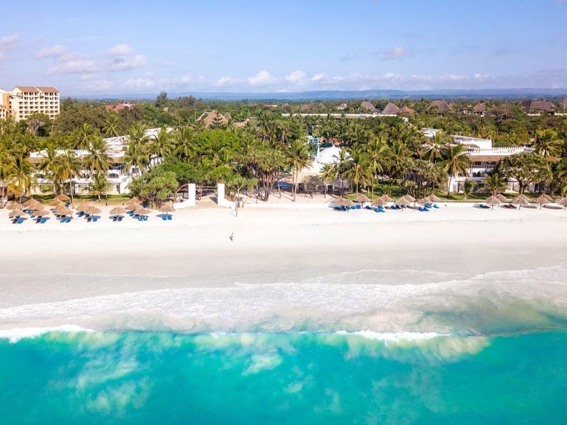 Bílá písečná pláž s tyrkysovou vodou a palmami, za nimi hotelové budovy a lehátka.