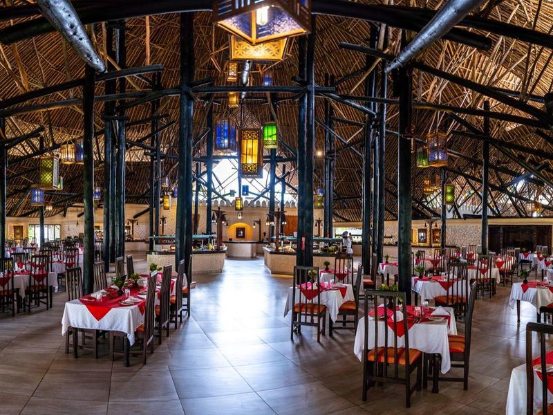 Velká restaurace s vysokými dřevěnými trámovými stropy, mnoha stoly a židlemi, teplé osvětlení.