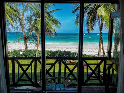 Blick durch eine offene Terrassentür auf weißen Sandstrand, Palmen und blaues Meer bei klarem Himmel.