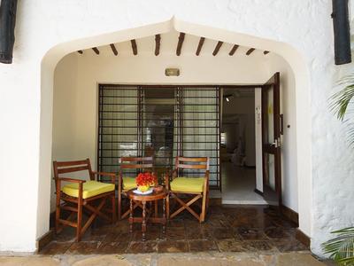 Veranda mit drei Holzstühlen und kleinem Tisch vor weißer Fassade, Sonnenschirm und Palme rechts.