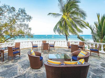 Offener Sitzbereich mit Stühlen und Tischen auf einer Terrasse mit Blick auf das Meer und Palmen.