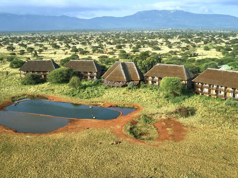 Lodge mit mehreren Hütten und einem Wasserloch in einer weiten grasbewachsenen Savannenlandschaft.