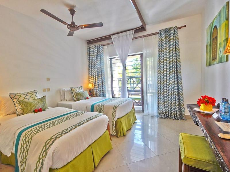 Helles Hotelzimmer mit zwei Betten, Lampen, Deckenventilator und Balkon mit Vorhängen.