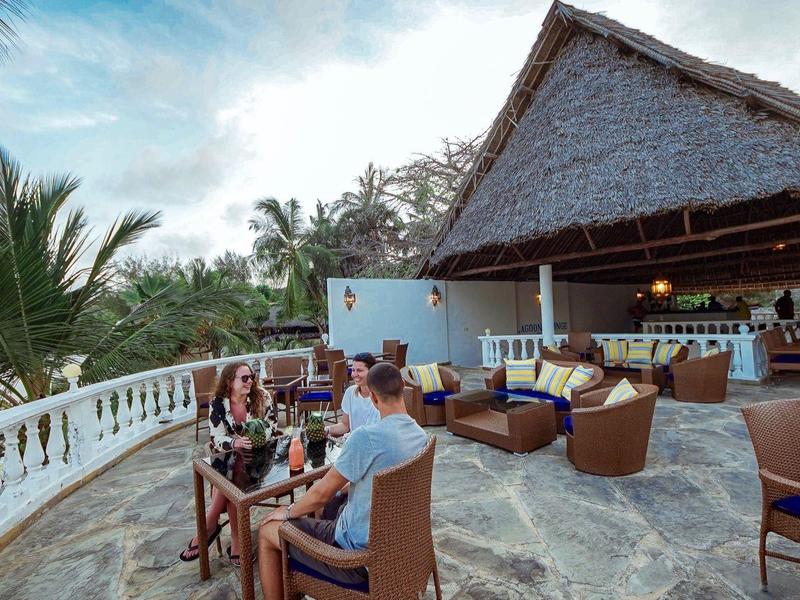 Terrassenbar mit Strohdach, Rattanmöbeln und Meerblick bei bewölktem Himmel.