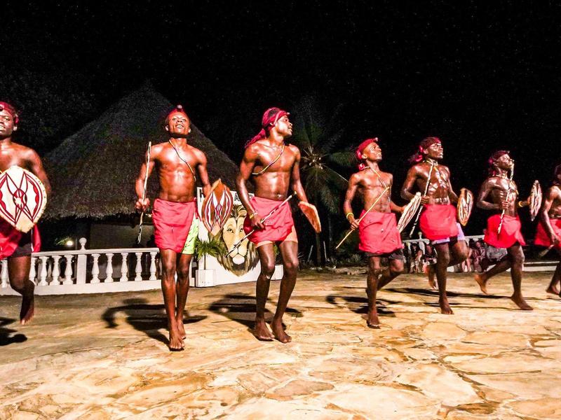 Tanzgruppe in traditioneller roter Kleidung tanzt nachts auf Steinboden vor dunklem Hintergrund.