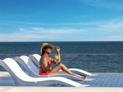 Frau in rotem Badeanzug mit Hut entspannt sich auf Liegestuhl am Pool mit Meerblick.