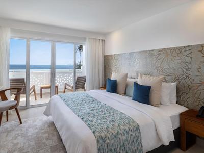 Helles Hotelzimmer mit Doppelbett, zwei Sesseln und Meerblick durch große Fenster.