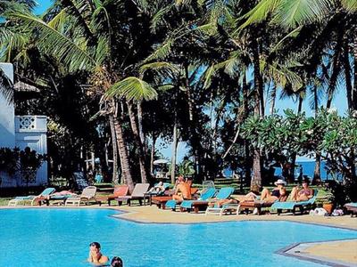 Des personnes nagent dans une grande piscine avec des chaises longues et des palmiers en arrière-plan.