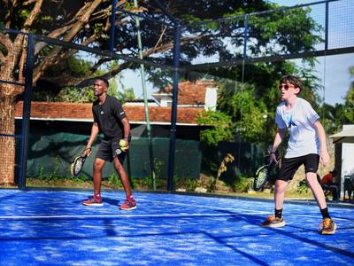 Zwei Männer spielen Padeltennis auf einem blauen Platz im Freien unter Bäumen.