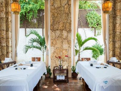 Deux tables de massage couvertes de blanc dans une pièce lumineuse avec des plantes et des murs en pierre.