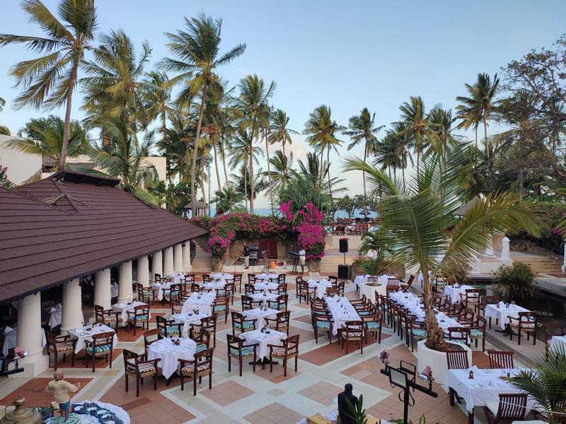 Espace repas extérieur d'hôtel avec de nombreuses tables dressées parmi les palmiers et plantes fleuries.