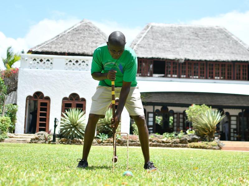 Homme jouant au golf sur la pelouse devant un grand hôtel tropical avec une véranda en bois.