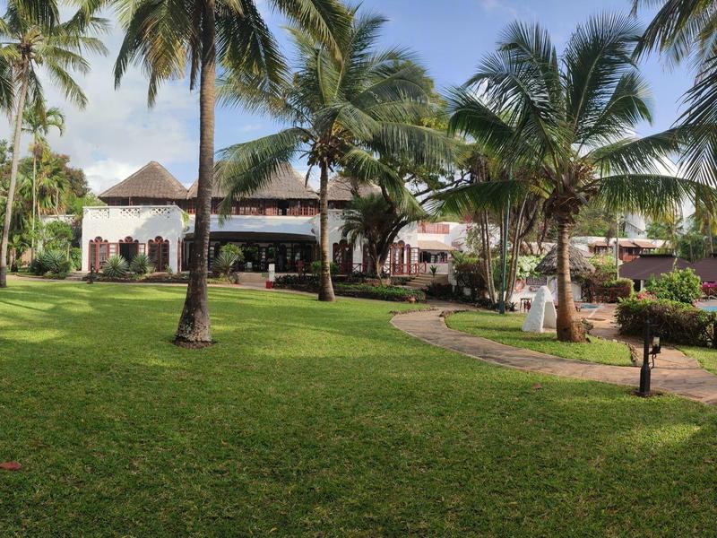 Jardin vert avec des palmiers et un sentier menant au bâtiment de l'hôtel dans un cadre tropical.