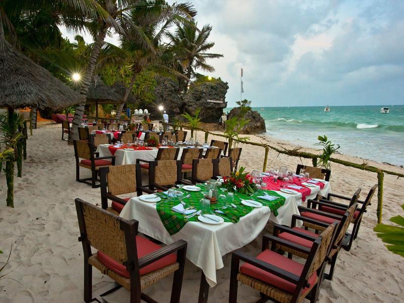 Restaurace s prostřenými stoly na pláži pod palmami a zataženou oblohou.