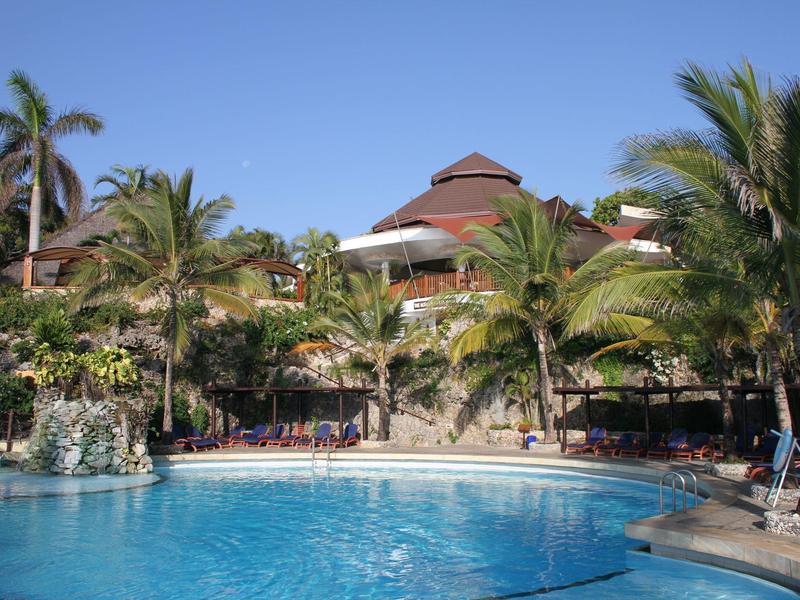 Luxusní hotel s velkým bazénem a palmami pod jasnou oblohou.