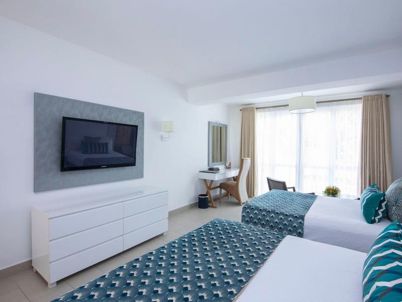 Světý hotelový pokoj se dvěma postelemi, televizí, psacím stolem a velkými okenicemi se závěsy.