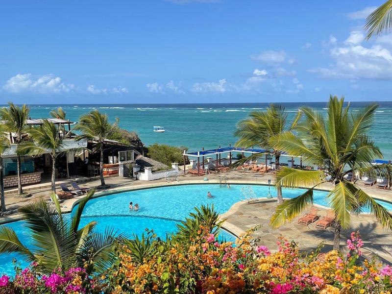 Hotelový bazén s palmami a výhledem na oceán pod modrou oblohou.