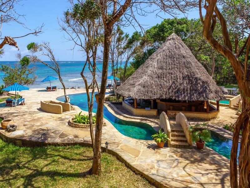 Station balnéaire avec bar au toit de chaume, piscine et vue sur la mer bleue parmi les arbres.