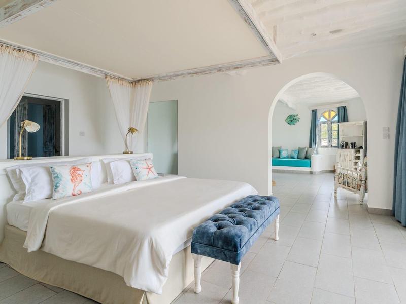 Chambre lumineuse avec grand lit, rideaux bleus et arche menant au salon.