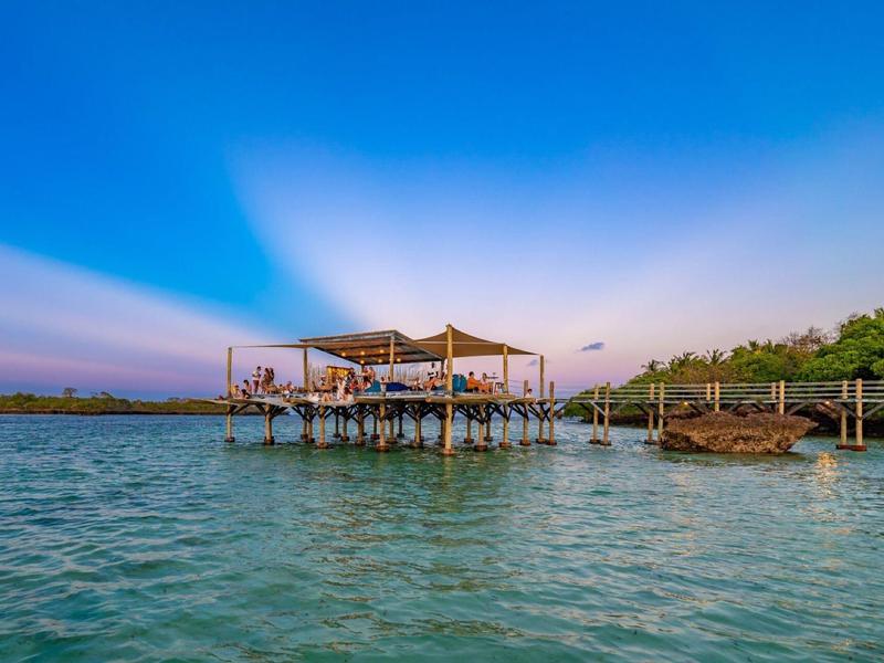 Un restaurant sur pilotis au-dessus d'une eau bleu clair avec un ciel nuageux en arrière-plan.