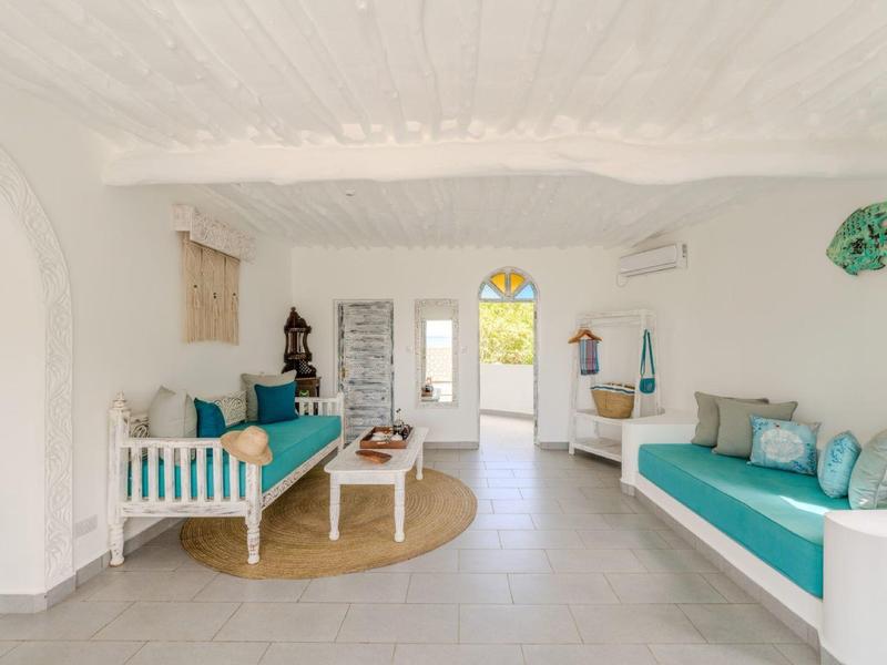 Salon lumineux avec murs blancs, coussins turquoise et tapis ronds dans un cadre de vacances.