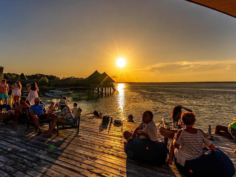 Des personnes profitent du coucher de soleil sur une terrasse en bois face à la plage.