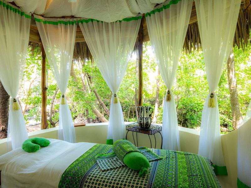 Chambre d'hôtel tropicale ouverte avec rideaux blancs et literie verte entourée de nature luxuriante.