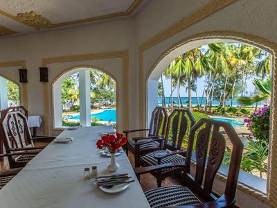 Zona pranzo con tavoli e sedie di legno, vista sulla piscina e palme attraverso grandi finestre.