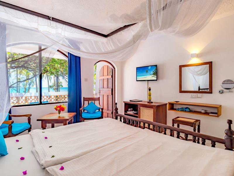 Camera d'hotel luminosa con letto matrimoniale, scrivania, TV e vista sul mare e palme.