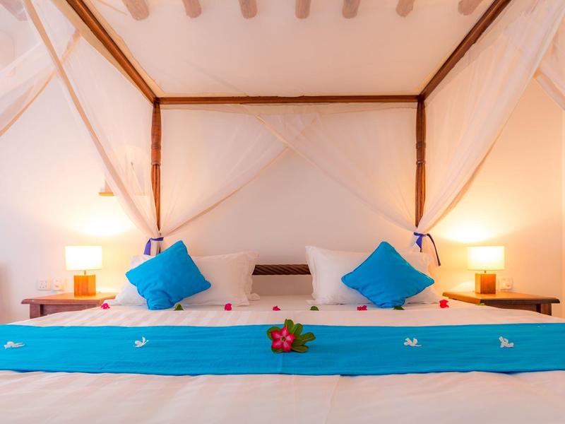 Un letto d'hotel con due cuscini blu, tende bianche e illuminazione calda ai lati.