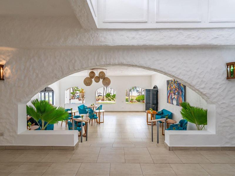 Area hotel luminosa con arco bianco, sedute blu e piante verdi sotto luce naturale.