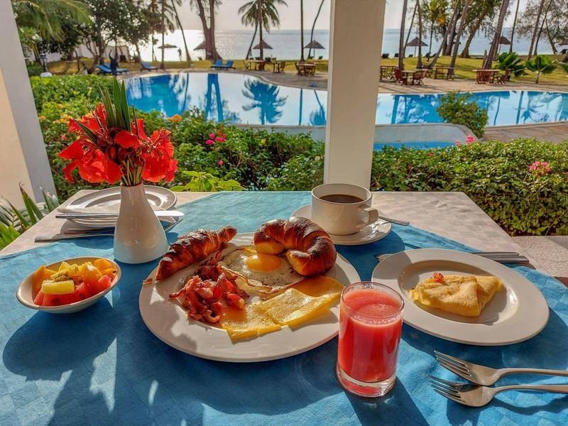 Un tavolo per la colazione apparecchiato con cibo e bevande con vista su piscina, palme e mare.
