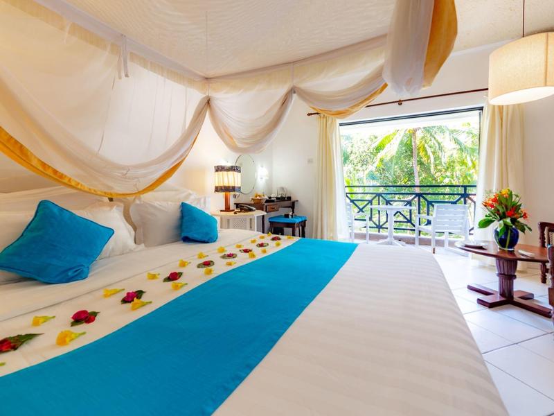 Licht hotelkamer met groot bed, kleurrijke kussens en balkon met uitzicht op planten.