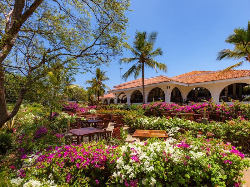 Hotel tuin met bloeiende struiken, palmbomen en een gebouw met bogen op de achtergrond.