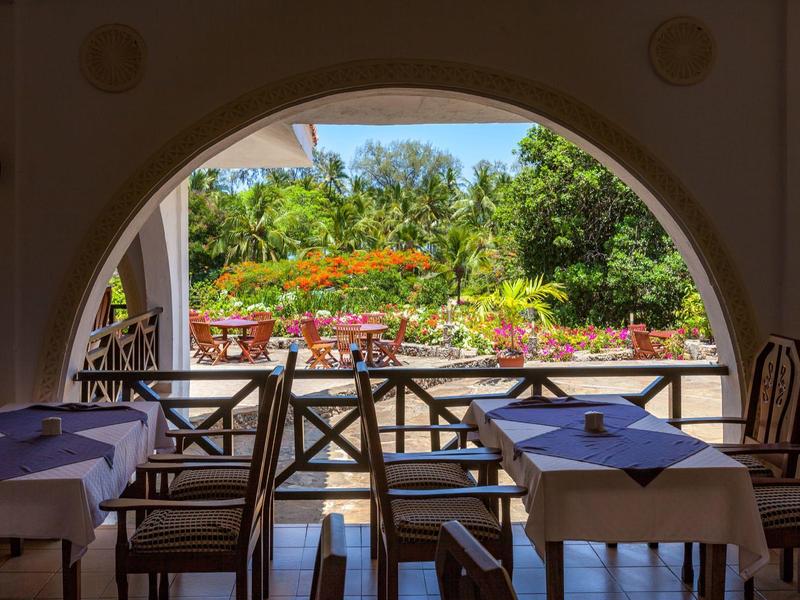 Uitzicht vanuit een schaduwrijk restaurant met gedekte tafels op een zonnige, bloeiende tuin.