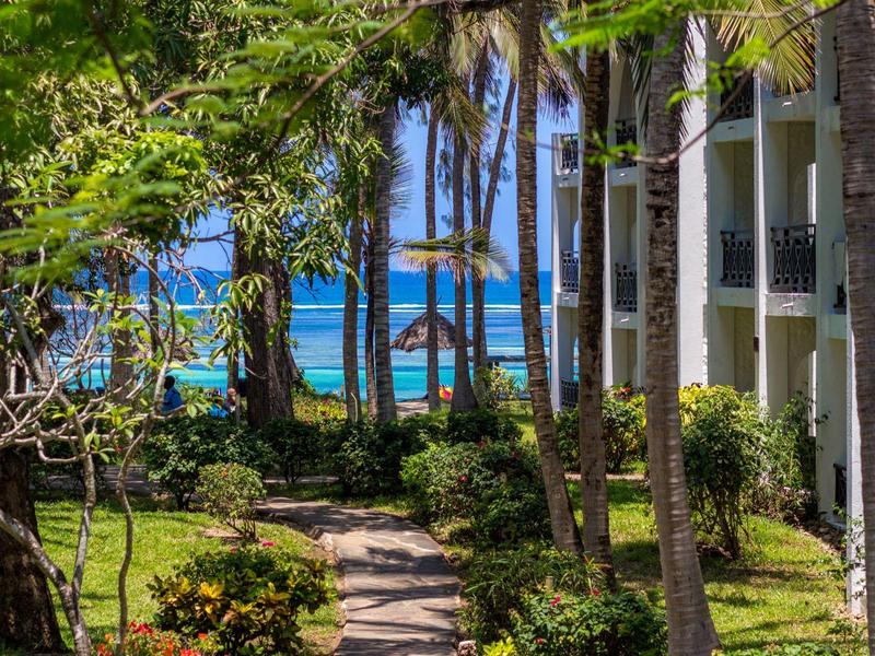 Uitzicht door tuin met palmbomen op strand en zee naast een hotelgebouw.