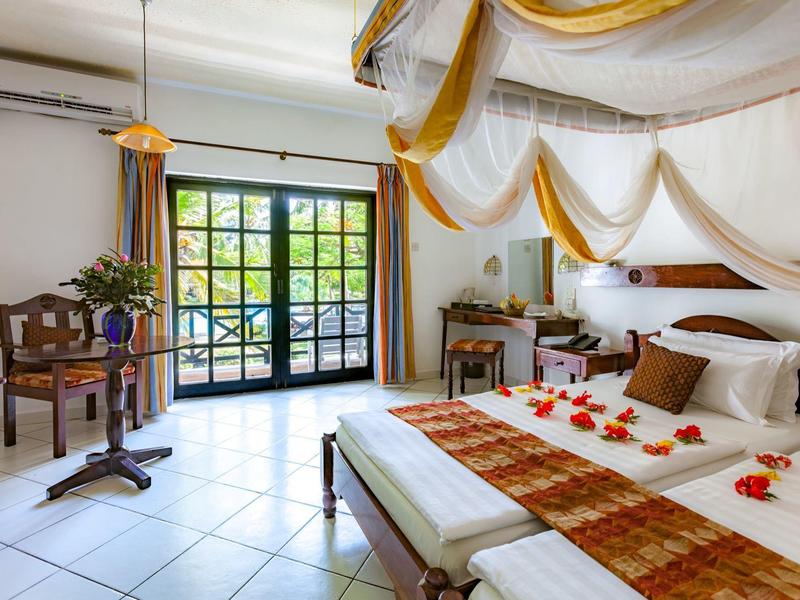 Lichte hotelkamer met groot raam, tweepersoonsbed met geel-witte versiering en bloemen.