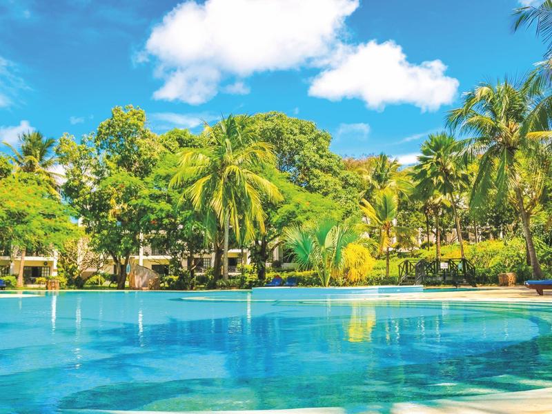 Groot zwembad omgeven door palmbomen en tropische begroeiing onder een blauwe lucht.