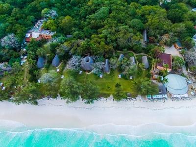 Luftaufnahme eines weißen Sandstrands mit grüner Vegetation und mehreren runden Hütten.