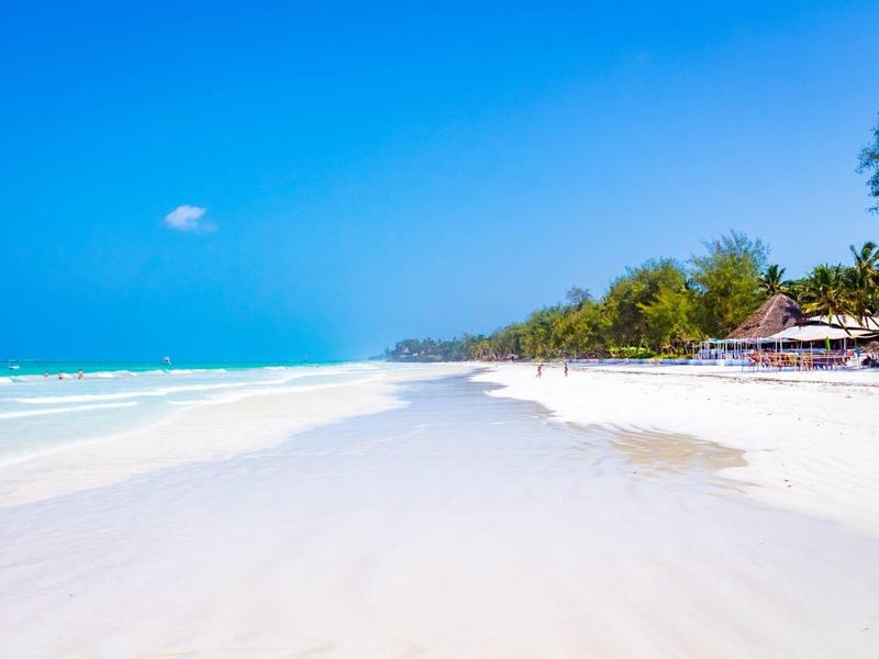 White sandy beach with clear blue sky and shallow sea, palm trees at the edge.