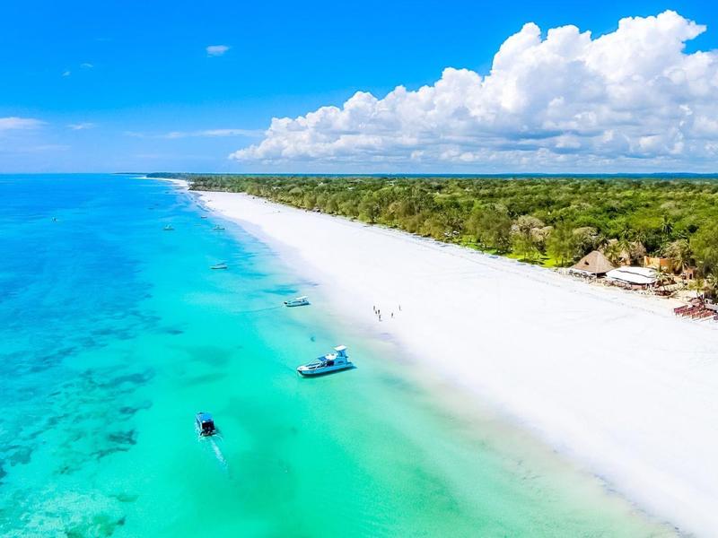 White sandy beach with clear turquoise water and boats under a sunny sky.
