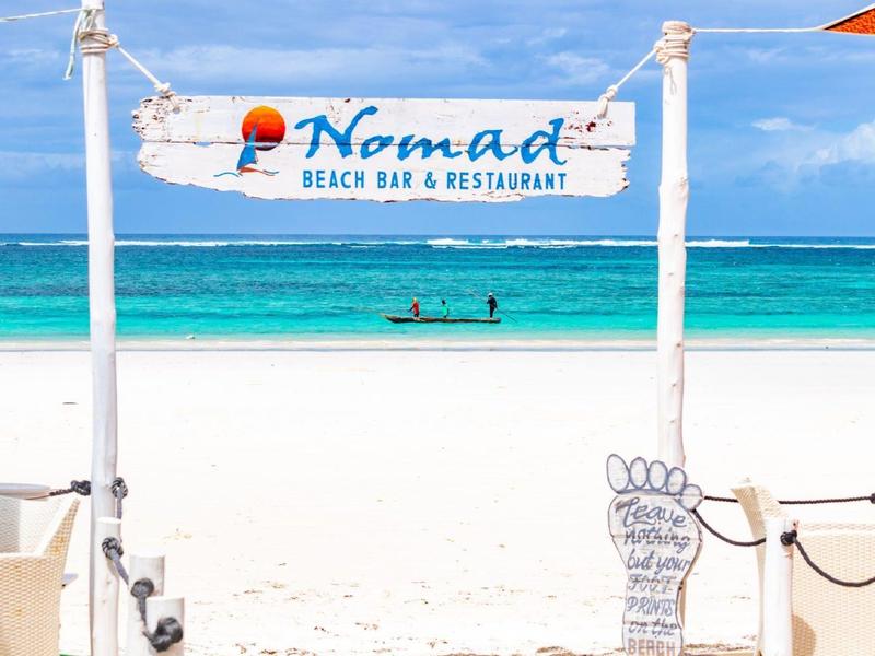 Beach with white sand and clear blue water, sign for Nomad Beach Bar & Restaurant.